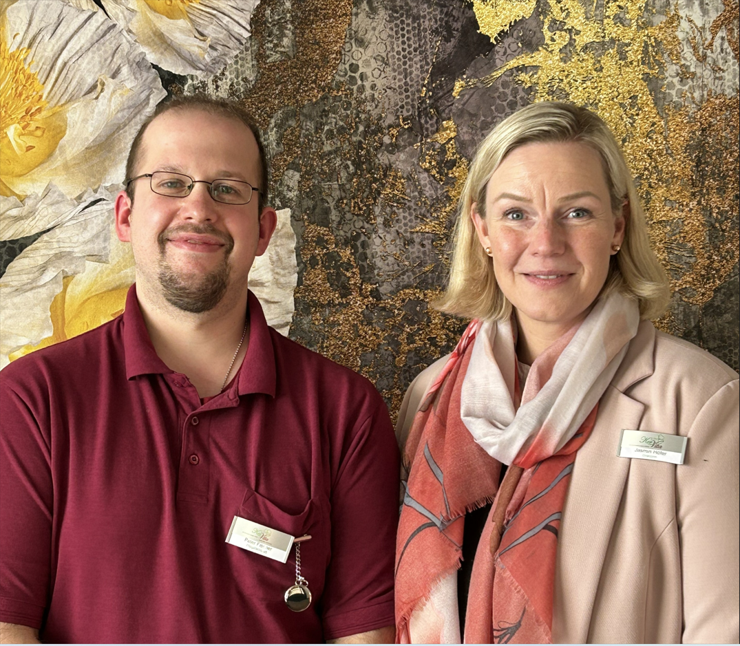 Ein Mann in bordeauxrotem Poloshirt und eine Frau in beigefarbener Jacke mit Schal stehen nebeneinander und lächeln in die Kamera. Beide tragen Namensschilder. Der Hintergrund zeigt eine künstlerisch gestaltete Wand mit floralen Elementen.